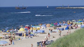 A beach with people sunbathing and swimming, boats in the water, and a lighthouse in the distance.