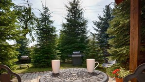 Une terrasse avec une table ronde et deux chaises, entourée de verdure et agrémentée d’un barbecue.