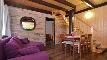 A cozy room with a purple sofa, wooden dining table, and chairs. A brick wall and wooden beams are visible.