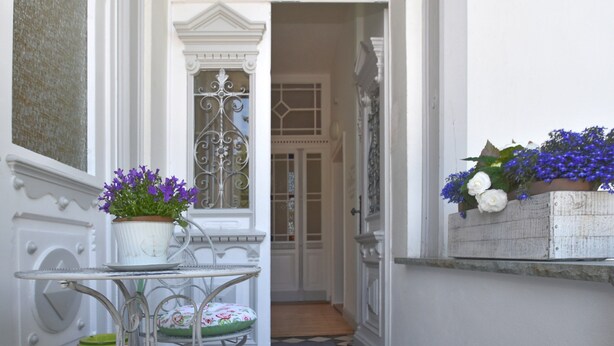 Une étroite entrée avec une porte blanche, une petite table avec une chaise, et des plantes en pot ornées de fleurs violettes.