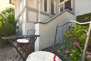 A patio with a metal table and chairs, a white cushion with red polka dots, and a flower box with blue flowers on the building's exterior wall. - Komfort mit Schlossblick (Blankenburg)