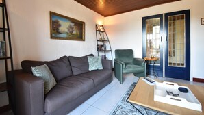 Ein Wohnzimmer mit Sofa, Sessel, Couchtisch, Bücherregal, Gemälde und einer Tür mit Glasverkleidung.