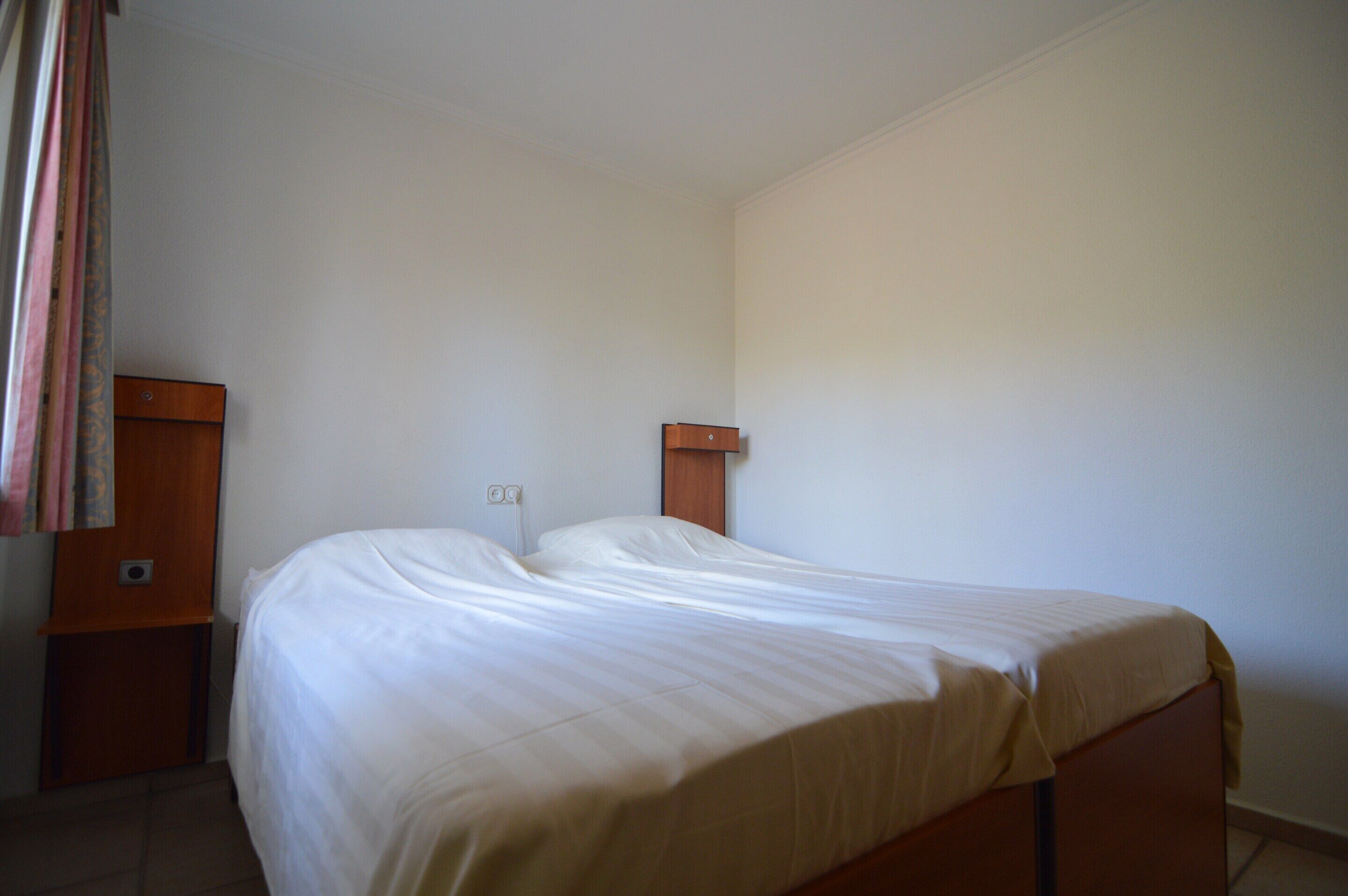 Een eenvoudige, spartanische slaapkamer met een enkel bed, nachtkastje en een houten hoofdbord.
