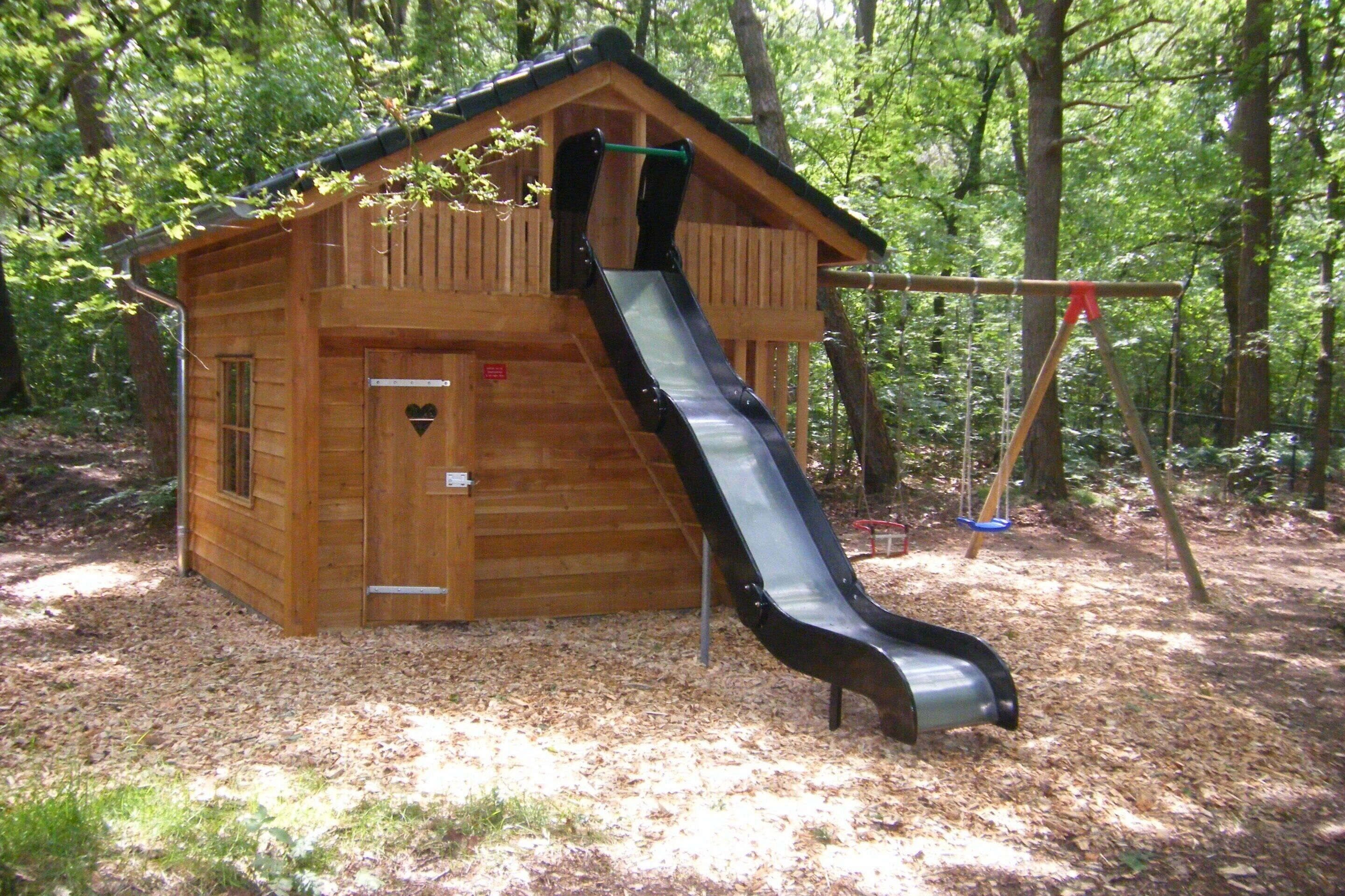 Een houten speelhuisje met een glijbaan en een deur in de vorm van een hart.