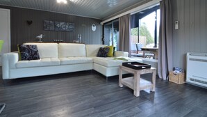A modern living room with a sectional sofa, a small wooden coffee table, and a large sliding glass door leading to an outdoor area. - Modern Waterfront Chalet (Geel)
