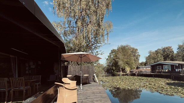 A wooden deck with outdoor seating and a table under a large umbrella, overlooking a calm water body with lily pads. - Modern Waterfront Chalet (Geel)