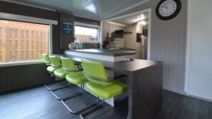 A modern kitchen with a bar area, green chairs, a clock, and a window with a view of a wooden fence. - Modern Waterfront Chalet (Geel)