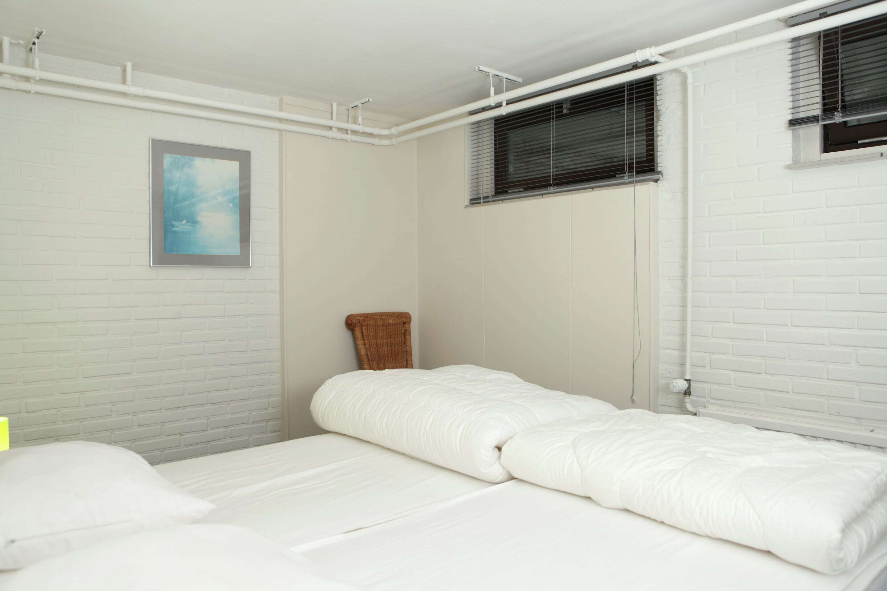 A room with a white brick wall, a framed picture, a window with blinds, and a bed with white bedding.