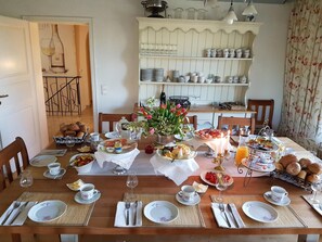 A dining table set with breakfast items, including bread, fruit, and beverages, in a room with a white cabinet and floral curtains. - Gästehaus Eschment (Bissersheim)