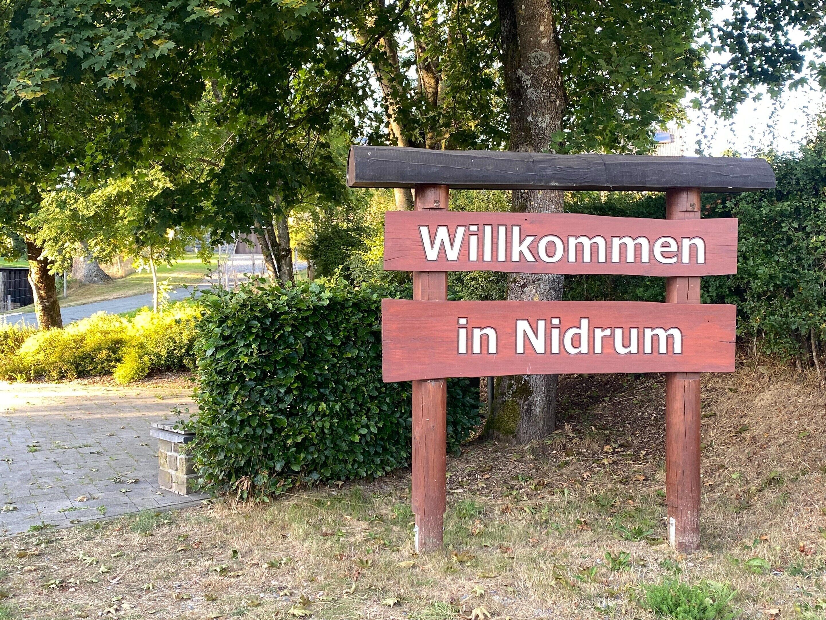 A wooden welcome sign with the text "Willkommen in Nidrum" in a natural setting with trees and a paved path.