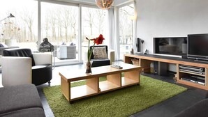 A modern living room with a large window, a flat-screen TV, a wooden coffee table, and a green rug. - Villa in Luttenberg Near National Park (Luttenberg)