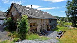 Une maison en pierre avec un toit en pente, une cheminée et une allée gravillonnée. On y trouve deux espaces de détente en plein air, avec des fauteuils et une petite table.