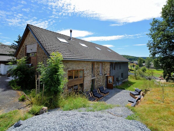 A stone house with a sloped roof, a chimney, and a gravel driveway. There are two outdoor seating areas with lounge chairs and a small table. - Modern Holiday Home in Meyerode With Terrace (Amel)