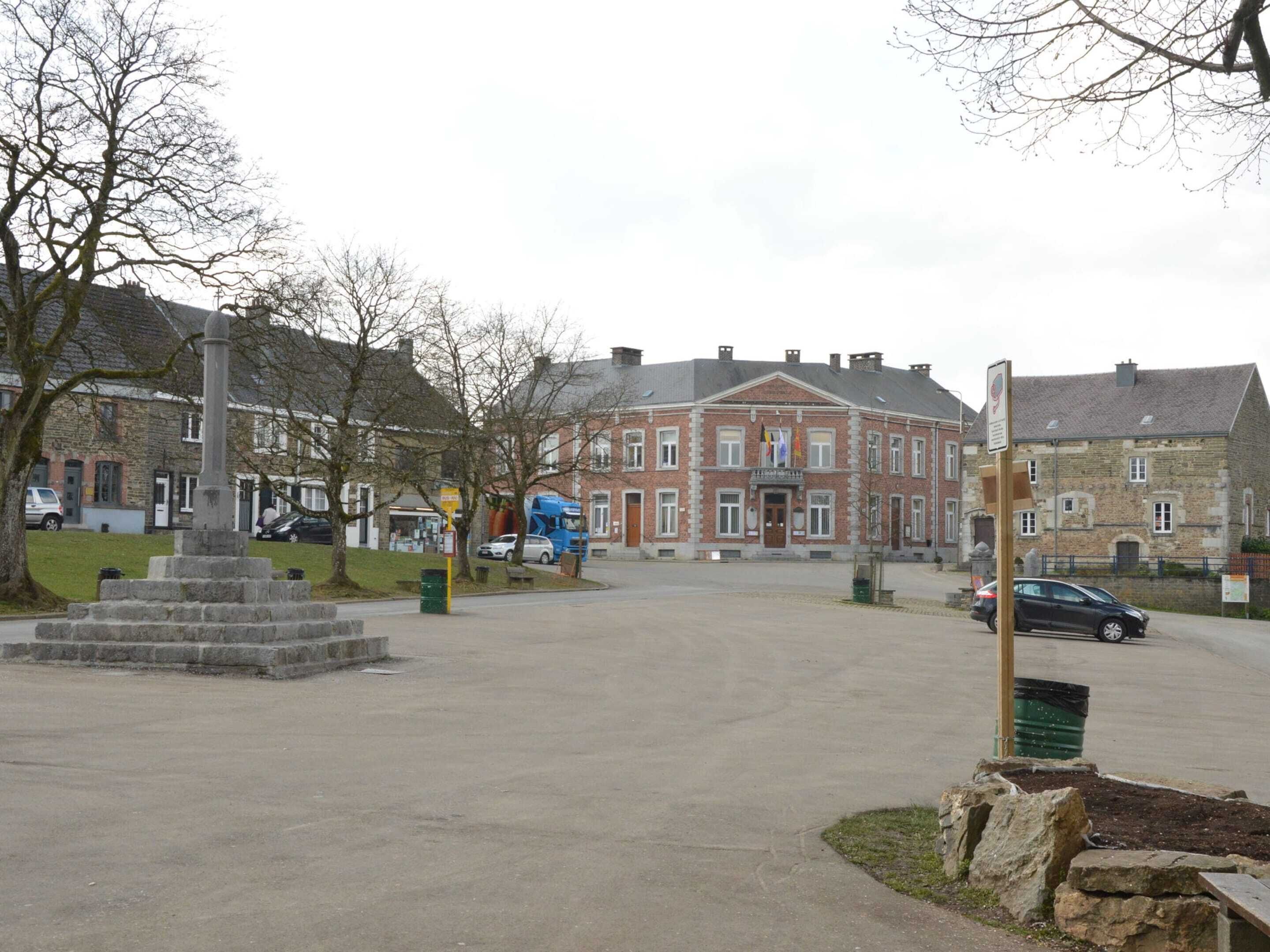 A public square with a stone monument, a red brick building, and a blue bus in the background.