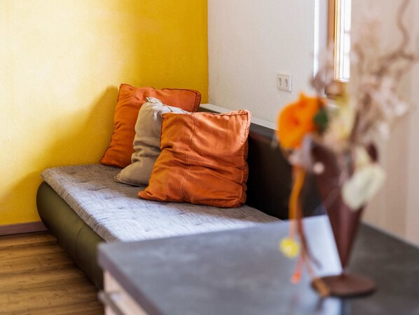 A small, cozy room with a sofa bed, orange and gray cushions, and a vase with flowers.