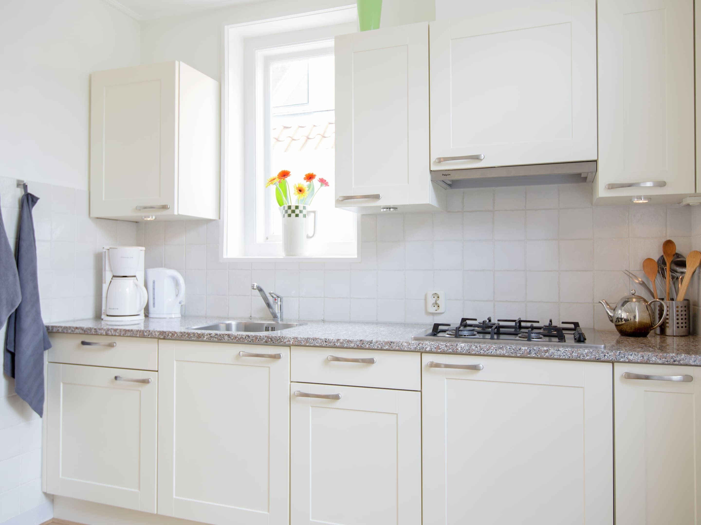 Una cocina con armarios blancos, una ventana con flores, una cafetera y una estufa a gas.