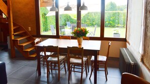 A dining area with a wooden table, chairs, and a vase of flowers. There is a staircase on the left and a view of a garden through large windows.