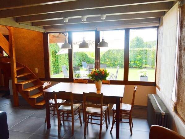 Een eetruimte met een houten tafel, stoelen en een vaas bloemen. Links bevindt zich een trap en er is uitzicht op een tuin door grote ramen.