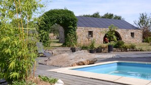 A swimming pool with a wooden deck, surrounded by greenery and a stone building with a green archway. - Charming Ardennes Getaway (Stoumont)