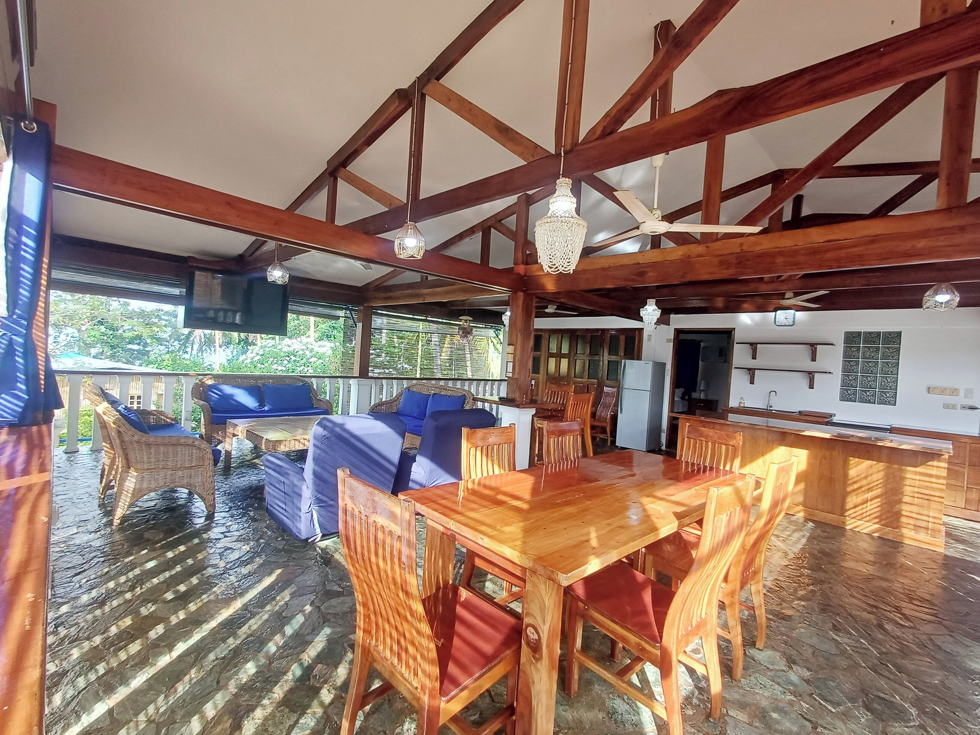 A wooden dining area with a table and chairs, a kitchen in the background, and a balcony with wicker furniture.