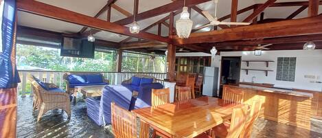 Un coin repas en bois avec une table et des chaises, une cuisine en arrière-plan et un balcon agrémenté de meubles en osier.