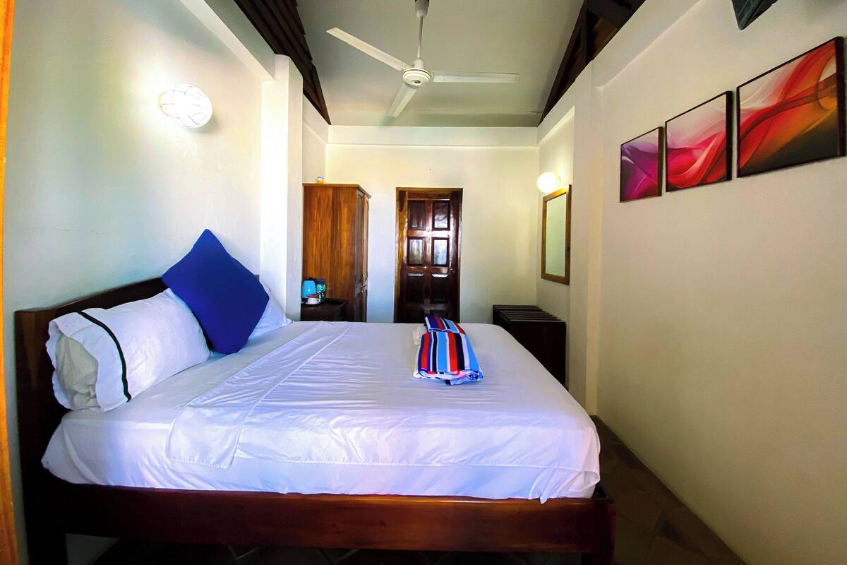 Une chambre d’hôtel comprenant un lit, un ventilateur de plafond, une table de chevet, une armoire et une porte donnant sur une autre pièce.