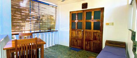 A room with a stone floor, a wooden table with chairs, a bed, a ceiling fan, and a window with blinds.