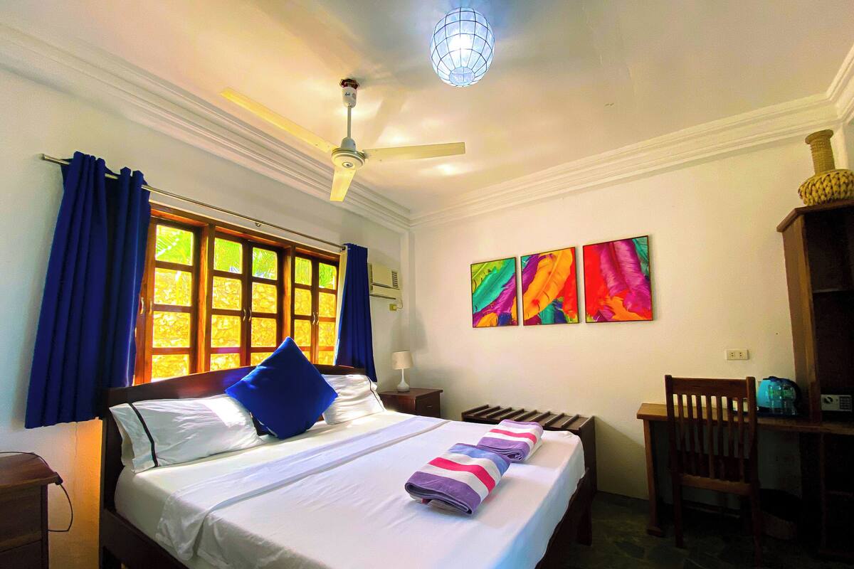 Une chambre d’hôtel équipée d’un lit, d’un bureau, d’une chaise, d’un ventilateur de plafond et d’une fenêtre avec des rideaux.