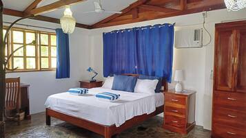 A bedroom with a wooden bed, blue curtains, a desk with a lamp, and a stone floor.