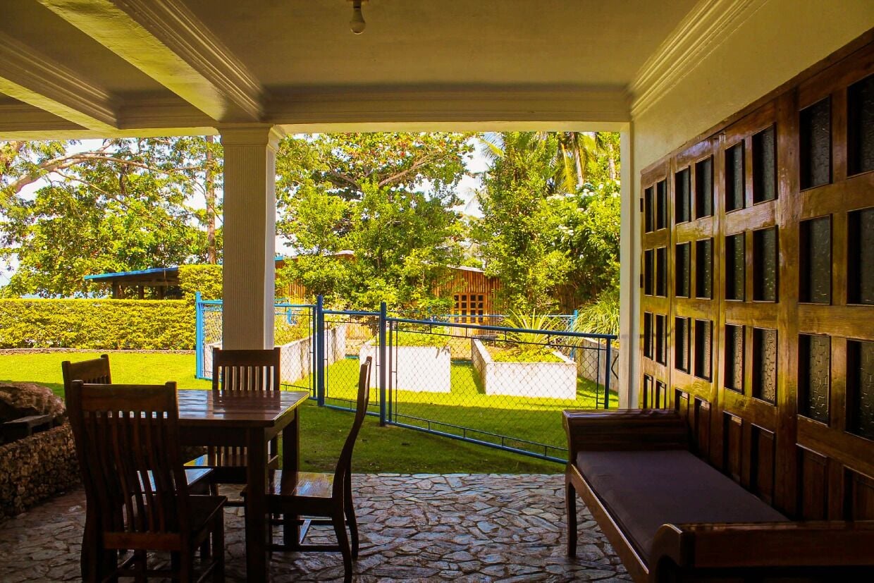 A covered patio with stone flooring, wooden furniture, and a view of a well-maintained garden and buildings in the background.
