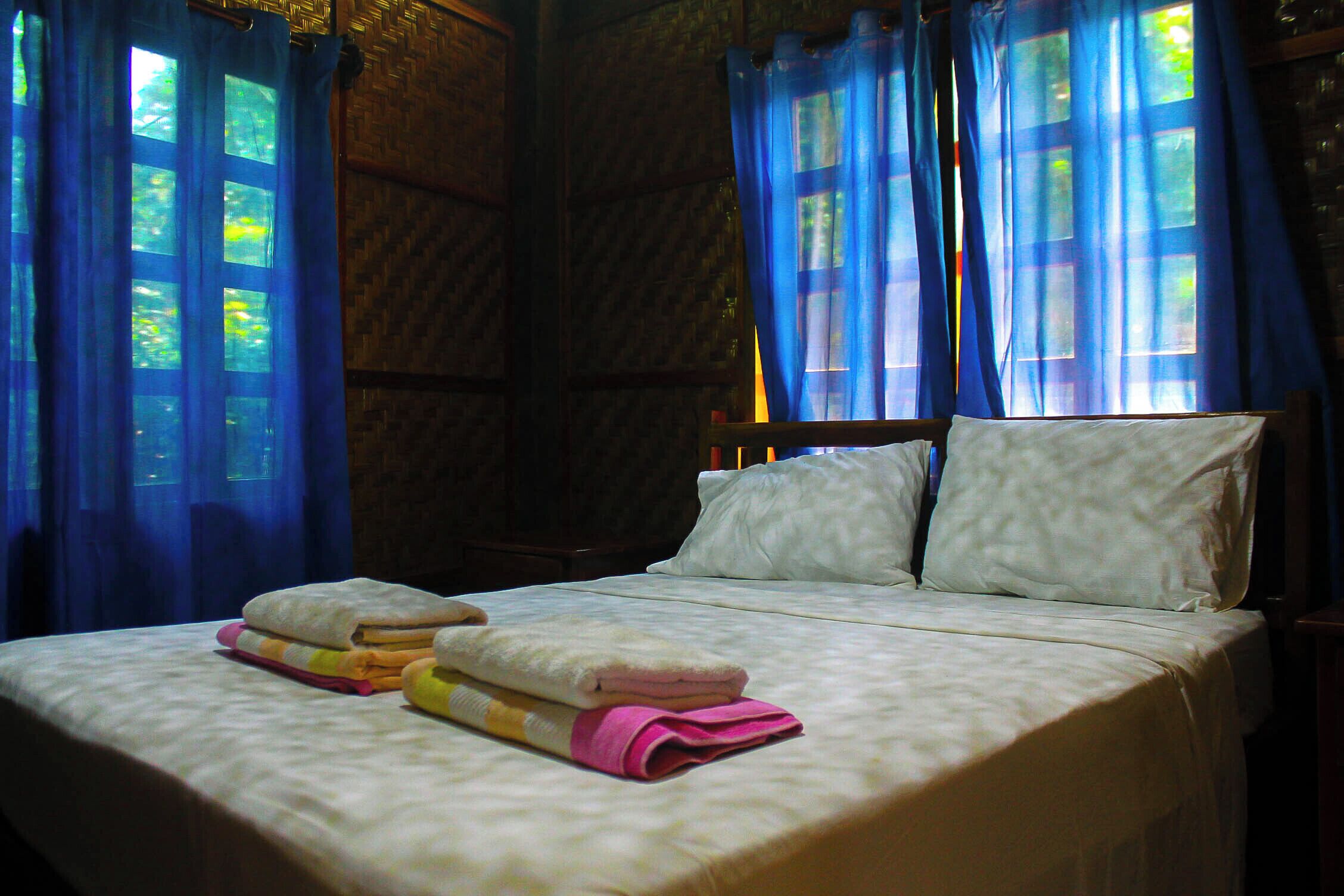 Un letto con lenzuola bianche, due cuscini e asciugamani piegati in una stanza con pareti in legno e tende blu.