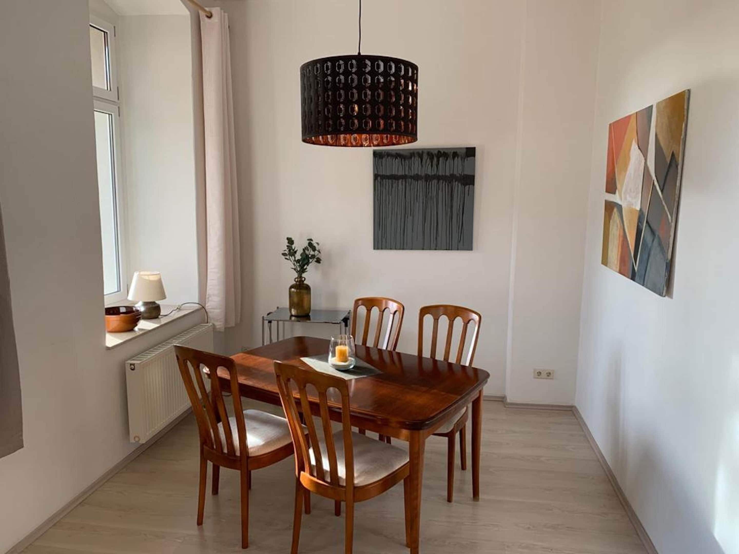 A dining area with a wooden table and chairs, a pendant light, a vase with a plant, and abstract art on the wall.
