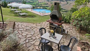 An outdoor patio with a stone table and chairs, a brick oven, and a pool in the background. - Apartment in Piticchio With Pool and BBQ (Arcevia)