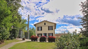 A two-story building with a garden in front, a metal gate, and a clear sky. - Apartment in Piticchio With Pool and BBQ (Arcevia)