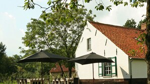 A white building with a red-tiled roof, a dining area with tables and chairs, and two large umbrellas. - Beernem Farmhouse With Pool (Beernem)