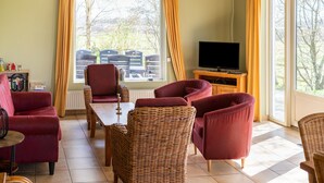 A living room with a television, wicker chairs, a red sofa, and a dining table. - Holiday Home with Meadow View near Forests (Schoonoord)