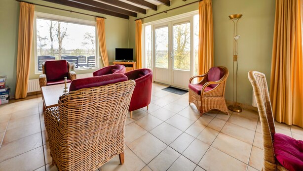 A room with a tiled floor, wicker furniture, a television, and large windows with curtains. - Holiday Home with Meadow View near Forests (Schoonoord)