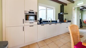 A kitchen with white cabinets, a built-in oven, a window, and a wicker chair. - Holiday Home with Meadow View near Forests (Schoonoord)