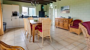 A kitchen with white cabinets, a dining table with wicker chairs, a wooden cabinet, and a red chair. - Holiday Home with Meadow View near Forests (Schoonoord)