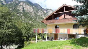 Eine zweistöckige Chalet mit Balkon, umgeben von üppiger Vegetation und Bergen im Hintergrund.