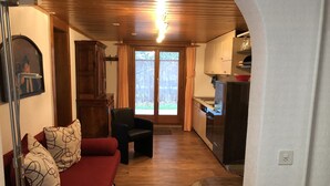 Ein gemütliches Wohnzimmer mit einem roten Sofa, einem schwarzen Sessel und einer Holzbalkendecke. Im Hintergrund ist eine Küche mit Herd und Schränken zu sehen.