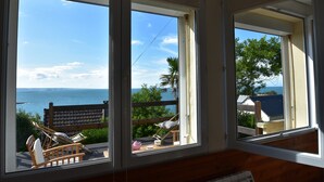 A room with a view of the sea, a wooden deck with chairs, and a window with curtains. - Sea View Stay in Fermanville (Fermanville)