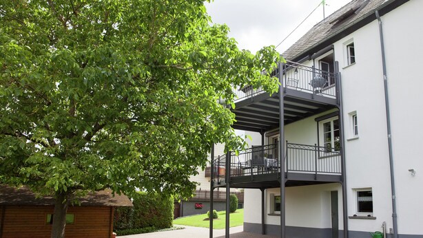 Un bâtiment à deux étages doté d’un balcon, avec un arbre devant et un chemin en briques.