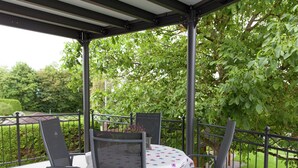 Une terrasse couverte, dotée d’une table et de chaises, entourée d’une rambarde en métal et de verdure.