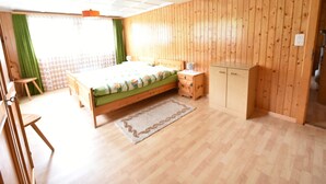 A bedroom with a wooden floor, a bed with a green bedspread, a nightstand with a lamp, a wooden wardrobe, and a window with green curtains. - Cozy Stay Near Aletsch (Grengiols)