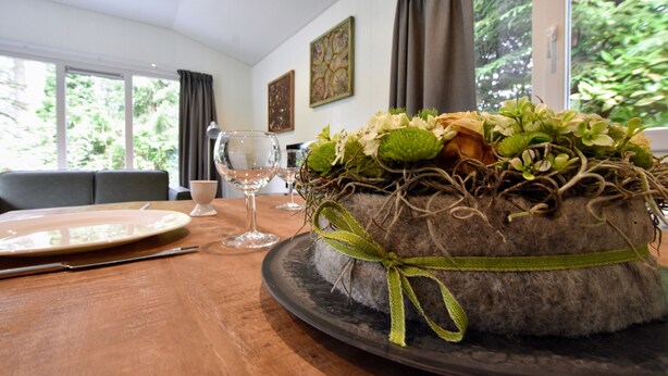 Un gâteau rond, orné d’un ruban vert, est posé sur une assiette sombre, elle-même placée sur une table en bois dans une pièce dotée d’une fenêtre et d’un tableau au mur.