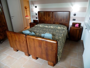 Un letto di legno con una coperta a motivi, un comodino con un fiore rosa e un punto luce a parete.