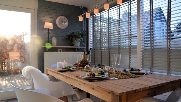 A dining table set with plates, glasses, and candles, surrounded by white chairs in a room with a stone wall, a clock, and a window with blinds. - Cozy Holiday Home in Noordwijkerhout near Lake (Noordwijkerhout)