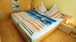 Een net opgemaakt bed met gestreept beddengoed en kussens, een nachtkastje met een lamp, en een houten garderobe.