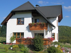 Ein zweistöckiges Haus mit geneigtem Dach, weißen Außenwänden und hölzernen Balkonen mit Blumenkästen.